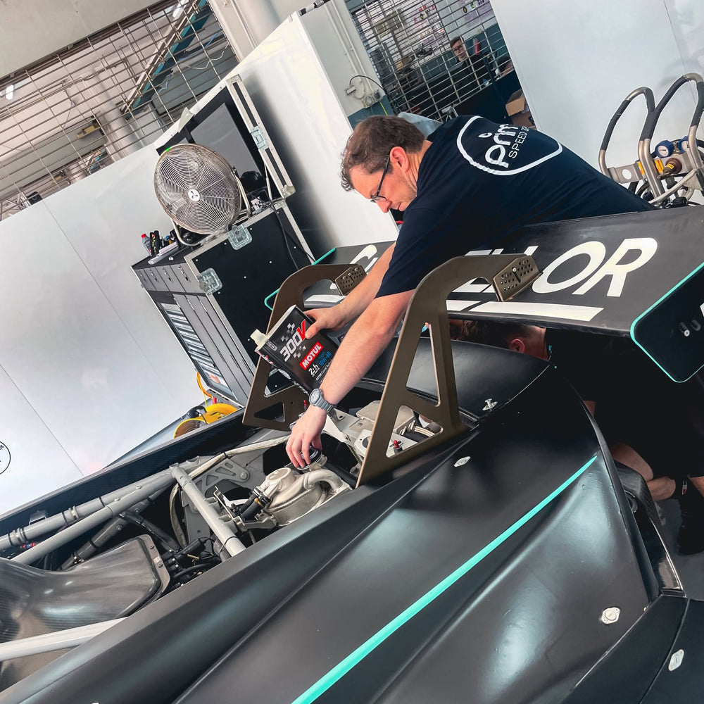 A man in a prime t-shirt works on a vehicle inside a pit garage. The vehicle is a matte black race car with a roll cage and teal accents.