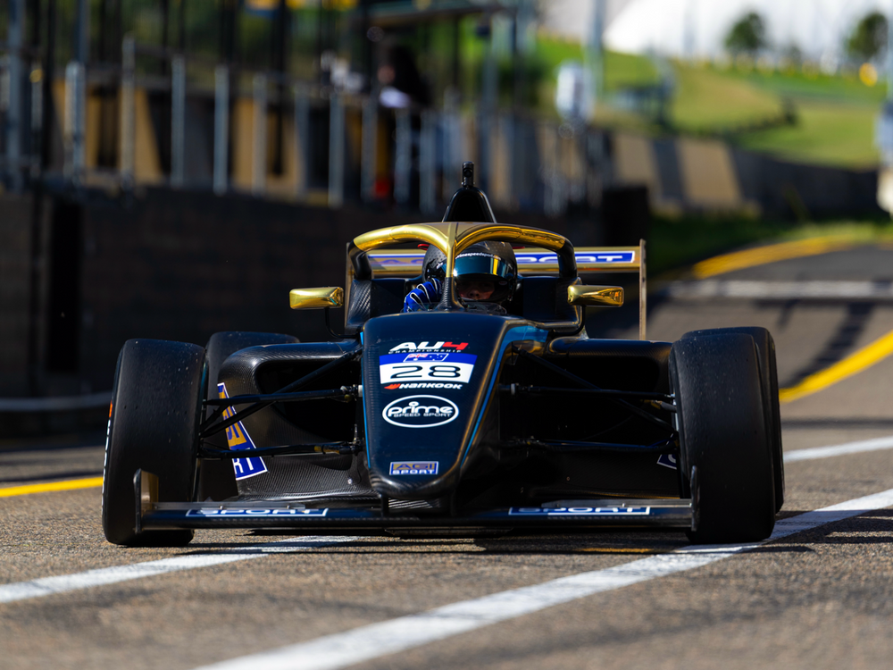 A black and gold f4 race car with sponsor logos, driven by a visible driver, racing on a track.