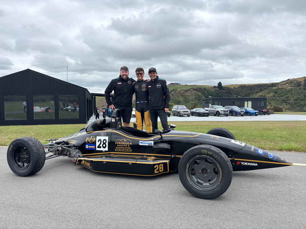 The Prime Speed Sport team standing with a black and gold Formula Ford race car (number 28) at a New Zealand racetrack under a cloudy sky.
