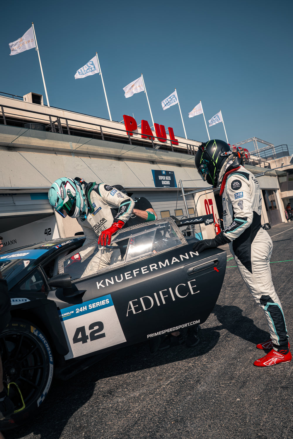 Two race car drivers standing next to a racing car on a track with flags in the background.