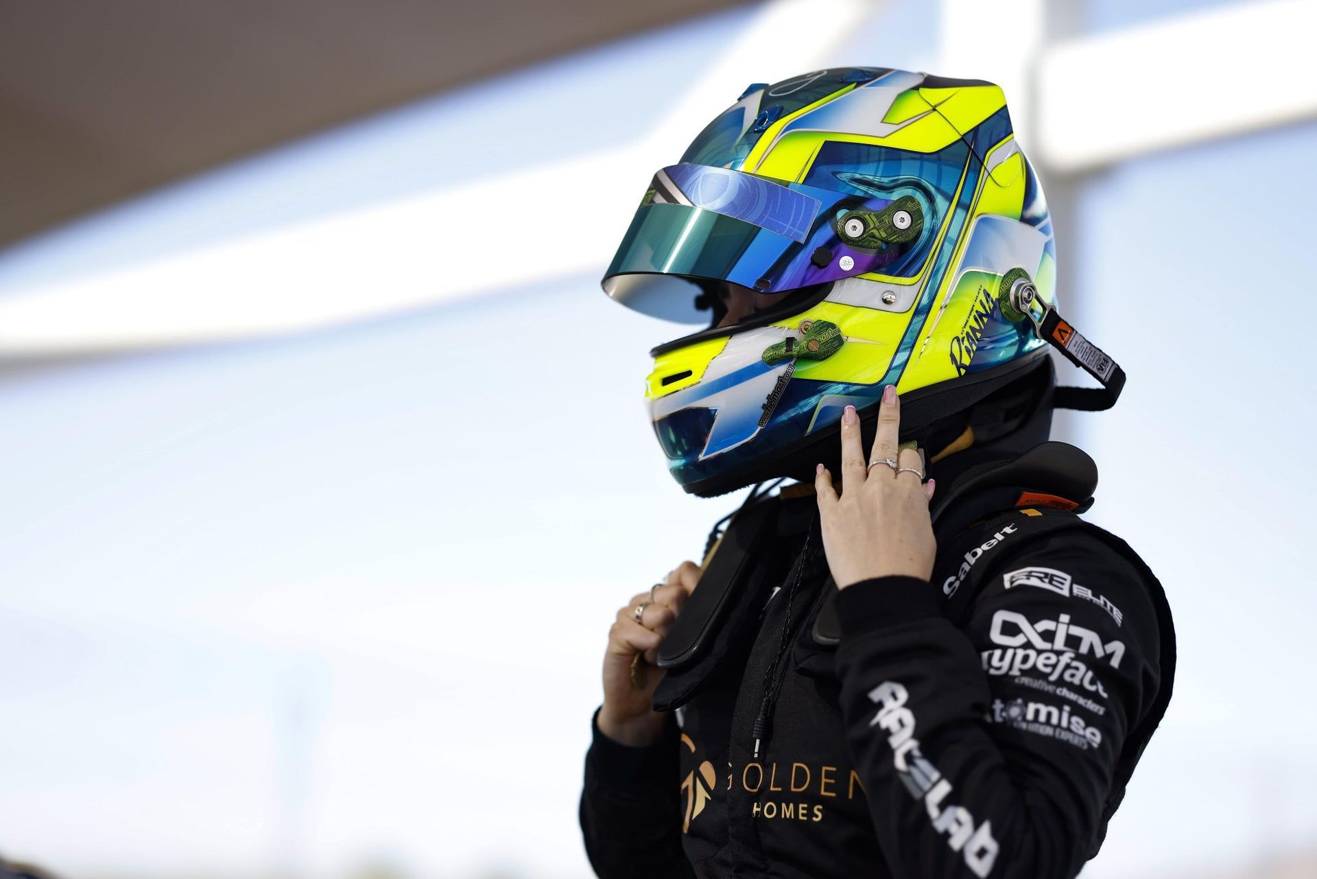 A girl in a black racing suit with sponsor logos and a colorful full-face racing helmet, standing against a bright outdoor background. Racing for Prime Speed Sport in a Porsche Cayman GT4
