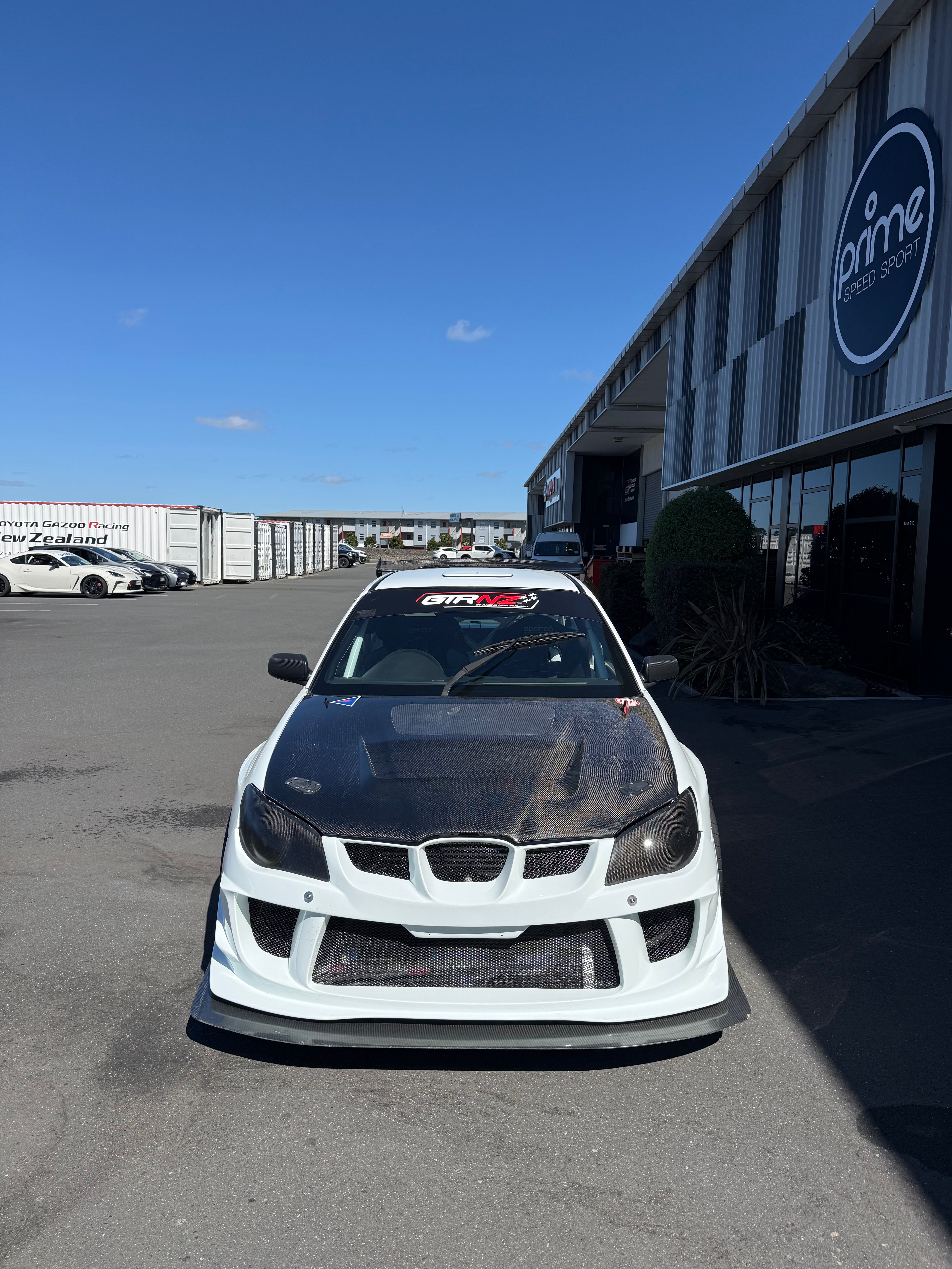 White Subaru Impreza car with a Carbon Fibre hood in front of a building with a logo on a clear day.